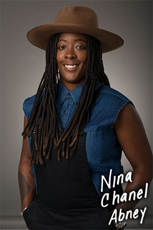 A portrait of a woman wearing a denim shirt, overalls and a brown hat in front of a grey background.